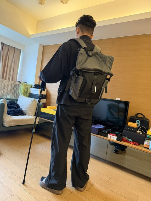 A man dressed in black stands in a living room wearing a gray tactical backpack and leaning on a black trekking pole.