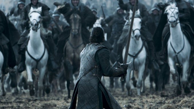 A scene from a medieval show featuring a figure in dark leather armor holding a sword, facing a charging group of horsemen in a grassy field.