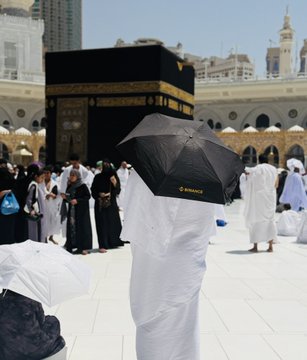 A person holds a black umbrella with the "Binance" logo near the Kaaba in Mecca. The image is tagged with "@binance", suggesting a connection or partnership.