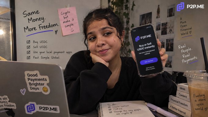 A woman sits at a desk holding a smartphone displaying the P2P.ME app for buying and selling USDC, surrounded by handwritten notes including 'Same Money. More Freedom.' and 'Good Payments Brighter Days' on a laptop sticker.