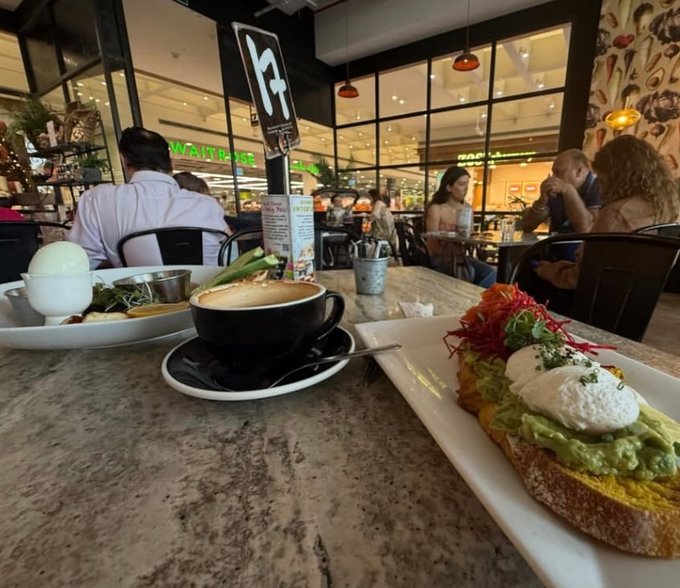 A first-person view of a meal at a cafe or restaurant, featuring avocado toast with a poached egg and microgreens, a coffee cup, and a boiled egg dish, with other diners and a 'WAITRESS' sign visible in the background.
