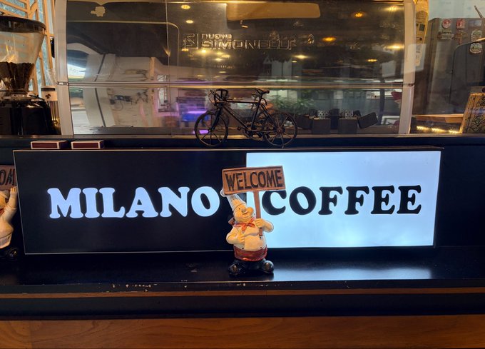 A storefront display featuring a lit sign reading "MILANO COFFEE" with a small chef figurine holding a "WELCOME" sign in the foreground.