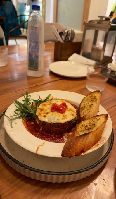 A plated dish of lasagna topped with melted cheese and a dollop of red sauce, served with garlic bread and arugula on a white plate, with a water bottle on the wooden table behind it.