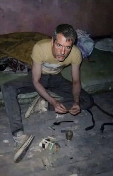 A man with gray hair sits on a dirt floor with various items scattered around him, including a pipe and what appears to be drug paraphernalia, in a dimly lit room with purple walls.