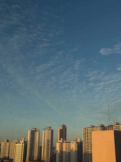 A cityscape photograph showing high-rise apartment buildings under a blue sky scattered with wispy clouds.