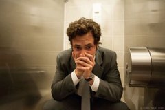 A man in a suit sits on a toilet in a public restroom, hands clasped over his face in apparent frustration or deep thought.