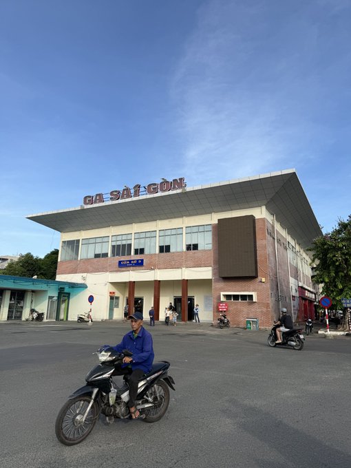 A man in a blue jacket sits on a motorbike in front of the "GA SÀI GÒN" (Saigon Station) railway building, with other motorbikes and pedestrians visible in the plaza. The image accompanies a casual Sunday greeting post, with the busy street scene reflecting everyday Vietnamese life and the hardworking spirit the message alludes to.