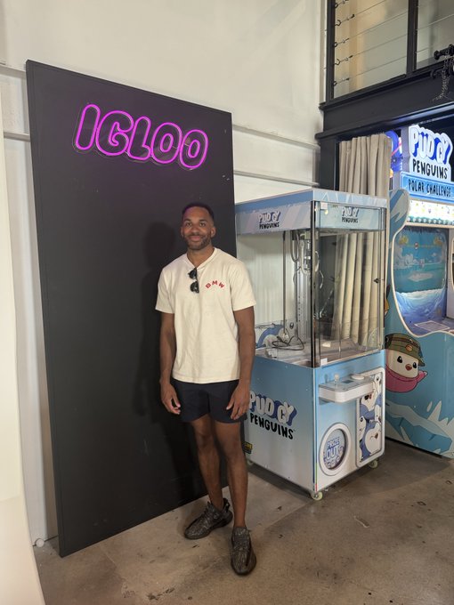 A man in a white t-shirt and dark shorts stands smiling between a black panel displaying a pink "IGLOO" neon sign and a blue "PUDGY PENGUINS" claw machine, fitting perfectly with the "gm especially to those who like penguins" vibe.