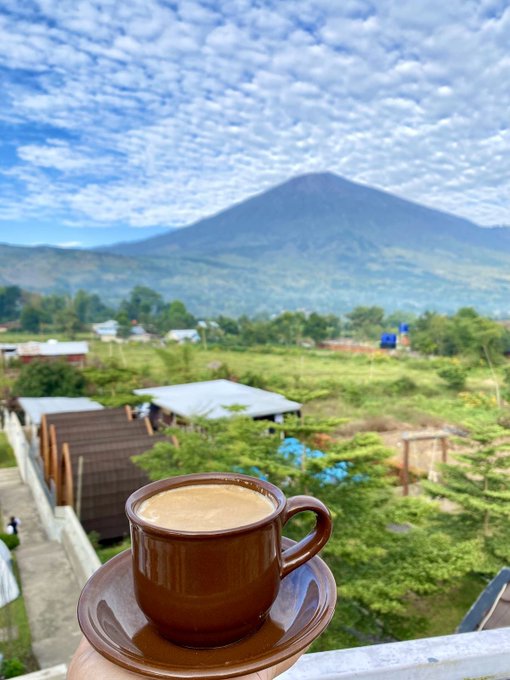 A hand holds up a brown cup of coffee, with a stunning landscape in the background featuring a large mountain under a sky filled with patterned clouds.