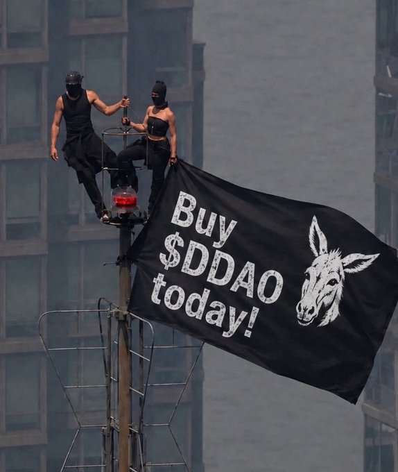 Two masked figures in black clothing stand atop a tall pole or structure, holding a large black banner that reads 'Buy $DDAO today!' featuring an illustration of a donkey's head.
