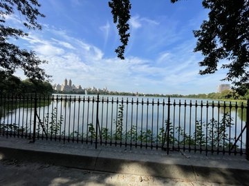 A serene view of the Jacqueline Kennedy Onassis Reservoir in Central Park, with the Manhattan skyline visible across the water, framed by a black iron fence and lush greenery. The peaceful park setting offers a quiet contrast to the busy Bitcoin Treasury meetings happening in New York.