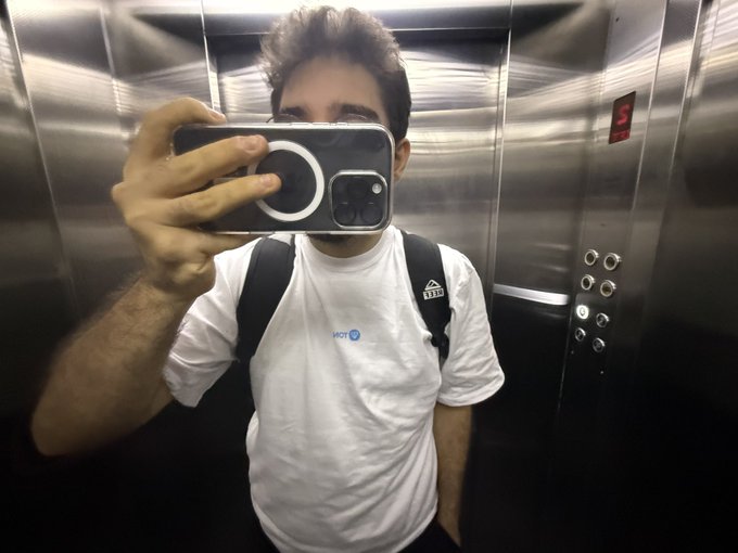 A man in a white t-shirt with a '1130' logo and a black backpack takes a mirror selfie with a smartphone in a metallic elevator.