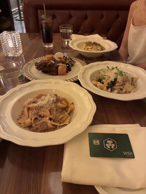 A photograph of a restaurant table set with four pasta dishes in white bowls, a glass of cola, water glasses, and a green Visa credit card resting on a napkin next to a person's arm.