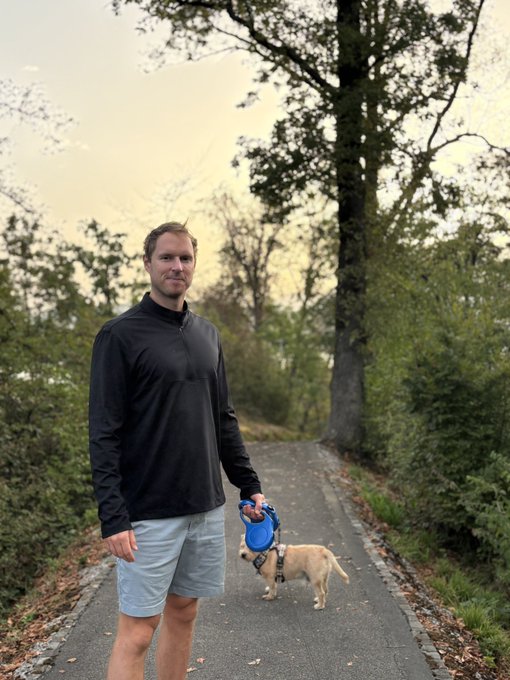 A man in a black long-sleeve shirt and grey shorts stands on a tree-lined path at sunrise, holding a blue retractable leash attached to a light-colored dog who is sniffing the ground. The peaceful morning walk with his furry companion reflects the post's themes of returning to a favorite sunrise spot and starting the day with positive energy.