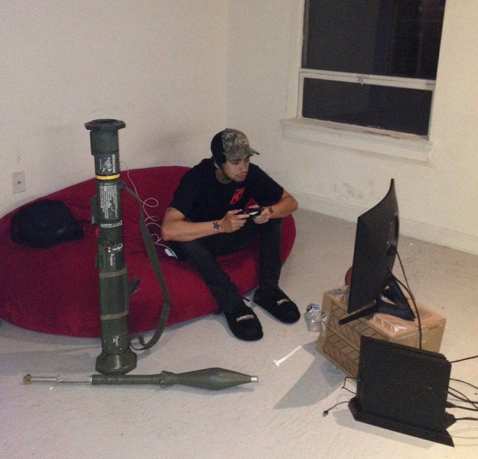 A young man in a black t-shirt, camo cap, and slippers sits on a red bean bag playing a video game, with a large green rocket-propelled grenade (RPG) weapon propped up next to him and a small monitor on a wooden crate serving as the screen.