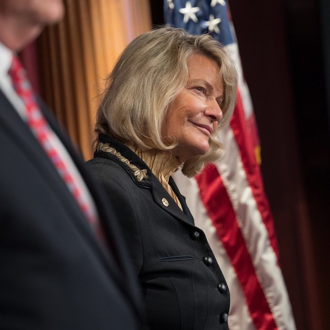 Senator Cynthia Lummis, wearing a dark blazer with gold embellishments, stands smiling before an American flag, apparently following remarks in which she accused Democrats of prioritizing their opposition to President Trump over advancing the CLARITY Act before the midterms.