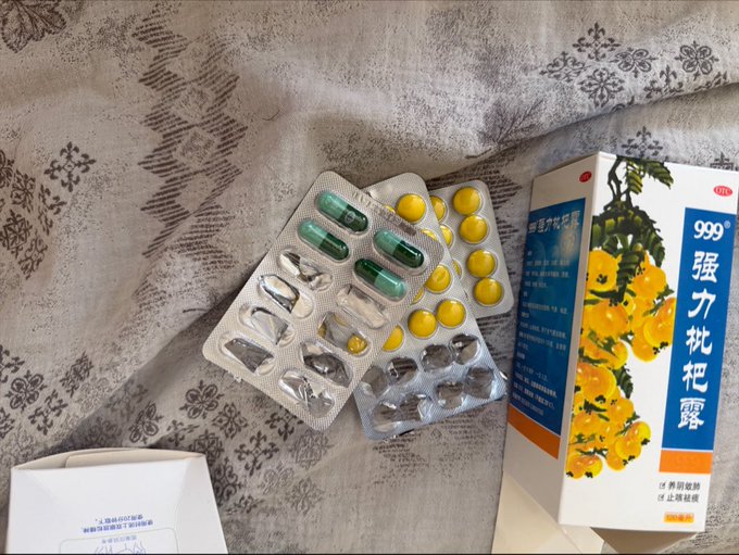 The image shows various blister packs of pills and capsules scattered on a patterned blanket next to a box labeled "强力枇杷露" (Strong Loquat Syrup) featuring loquat fruit imagery and the number "999." The scene reflects the post's theme of feeling unwell—struggling with a sore throat and eye pain—while the user humorously debates which medicines to take, seemingly lost in self-medication.