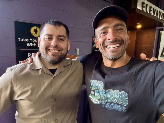 A selfie of two smiling men; the one on the right wears a black t-shirt reading 'XRP VEGAS 2026'.