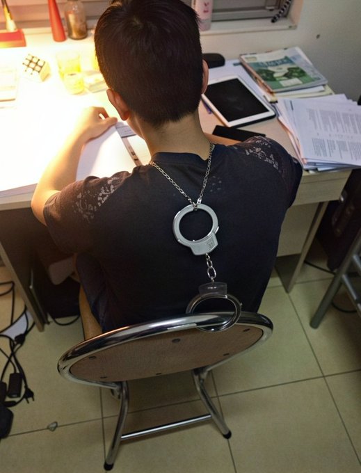 A woman sits at a desk working, wearing a black top and lace shawl, with handcuffs attached to a chain around her neck and cuffed to the back of a wooden chair.