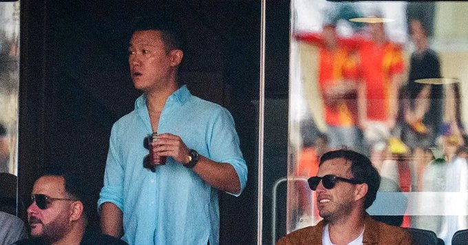 A man in a light blue shirt holds a drink while standing in what appears to be a stadium viewing box, with other men wearing sunglasses seated nearby.