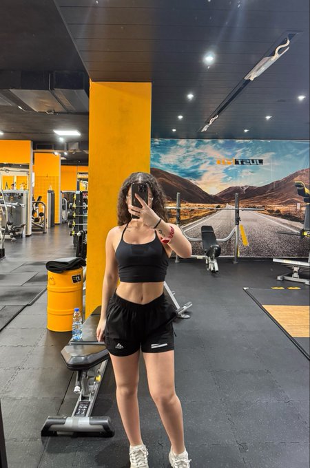 The image features a woman taking a selfie with her phone, standing in front of a gym. She appears to be posing for the camera while wearing black shorts and a tank top. The background includes various exercise equipment such as dumbbells and weight machines, indicating that she is at a fitness center or gym. The caption on the image reads "been more consistent since the start of this month", suggesting that the woman has been consistently working out and maintaining her fitness routine.