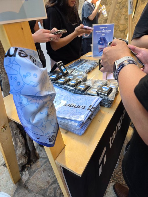 A vendor table displays stacks of 'tangent' branded blue bandanas, black cards, and small gadgets, with people using smartphones around it.