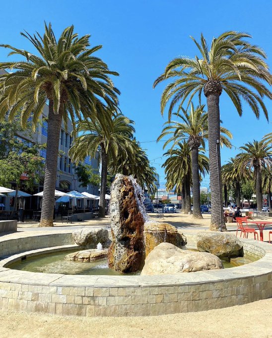 A photograph of a stone fountain with water flowing from a central rock formation, surrounded by tall palm trees in a sunny outdoor plaza setting.