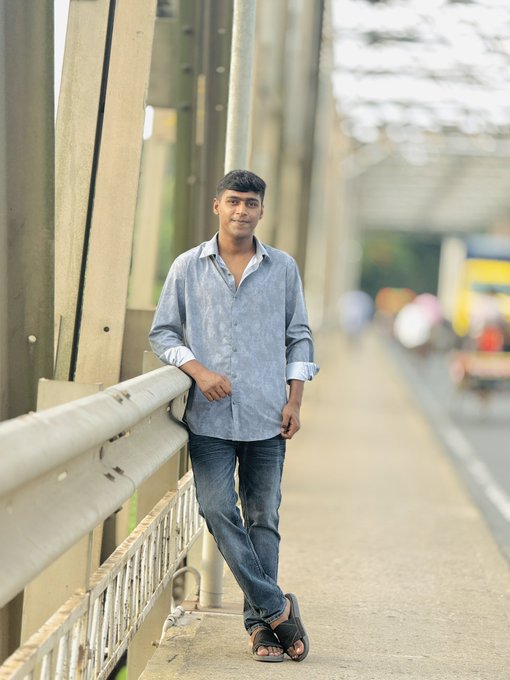A young man in a light blue button-up shirt and jeans leans casually against a bridge railing on a sunny day, smiling at the camera.