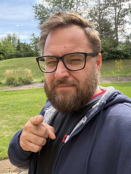 Outdoor selfie of a bearded man wearing black-rimmed glasses and a navy blue zip-up hoodie over a red shirt, pointing at the camera with a grassy park and trees in the background.