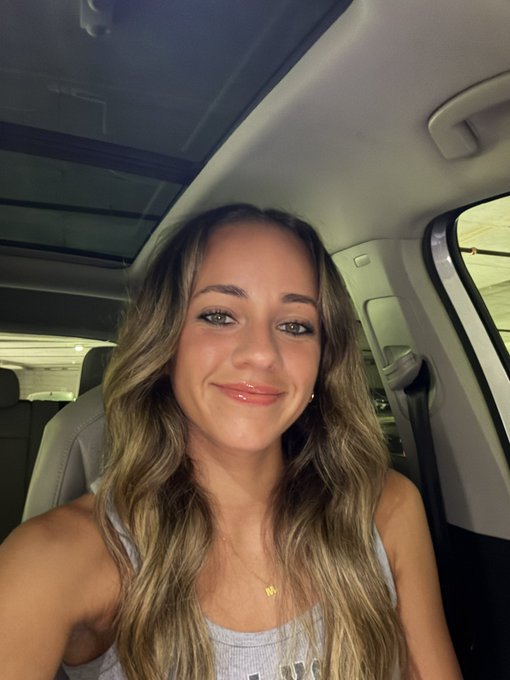 Close-up selfie of a young woman with long wavy highlighted blonde-brown hair, wearing a gray tank top and small star necklace, sitting inside a car.