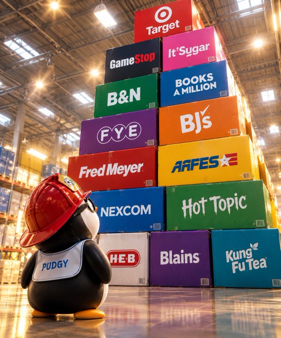 A penguin character wearing a red hard hat and a shirt labeled 'PUDGY' stands before a tall stack of colorful boxes in a warehouse, with boxes bearing retail names like 'Target', 'GameStop', 'B&N', and 'NEXCOM'.