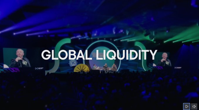 A large conference audience faces a stage illuminated by purple and green lighting, where the text "GLOBAL LIQUIDITY" is prominently displayed across the center, reinforcing the post's theme. Two large screens on either side show a man speaking into a microphone, connecting the live event to the discussion of global liquidity and XRP.