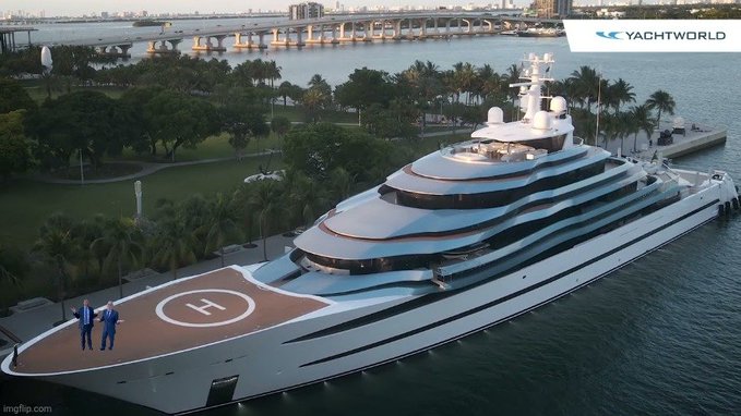 An aerial view of a large luxury yacht docked near a coastline with a helipad marked 'H' on the bow, branded with the 'Yachtworld' logo.