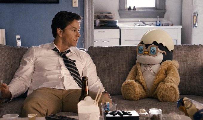 A scene from a film or TV show where a man in a white shirt and striped tie sits on a couch next to a large stuffed monkey mascot wearing an eggshell on its head and flower-shaped glasses.