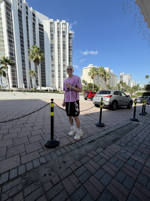 A man in a pink t-shirt and black shorts stands confidently on a sunny brick sidewalk in what appears to be Miami, with tall white high-rise buildings, palm trees, and parked cars in the background, embodying the post's upbeat "Good Morning" greeting to genuine connections while blocking out scammers, mirroring his own no-nonsense energy as he poses beside chain-link barriers.