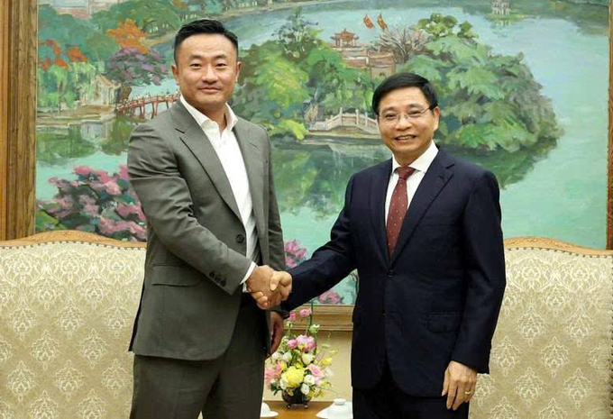 The image shows two men shaking hands, both dressed in suits with ties. They are standing next to each other near a painting of a lake surrounded by trees. The man on the left side of the image has his hand placed over the other man's shoulder, while the second man is facing away from the viewer and looking directly at him. Both men appear to be engaged in a formal handshake, possibly as part of an official or professional event.