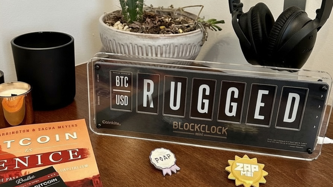 A desk setup featuring a Coinkite Blockclock mini displaying 'BTC USD RUGGED', alongside a 'Bitcoin & Venice' book by Riverton & Sacha Meyers, POAP and ZAP badges, headphones, and a potted plant.