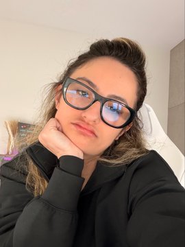 A casual selfie of a woman wearing black-rimmed glasses and a black sweatshirt, resting her chin on her hand with a slight smile.