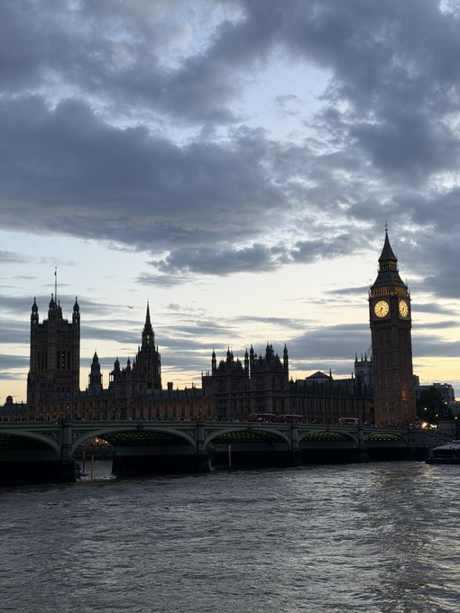 A scenic view of the Palace of Westminster and Big Ben at dusk, with Westminster Bridge spanning the Thames under a cloudy sky.