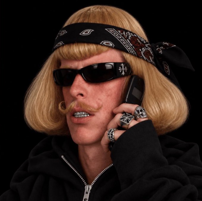 A man wearing a black bandana, dark sunglasses, a blonde bob wig, and a curled blonde mustache holds a flip phone to his ear while sporting multiple silver rings, including skull and cross designs.