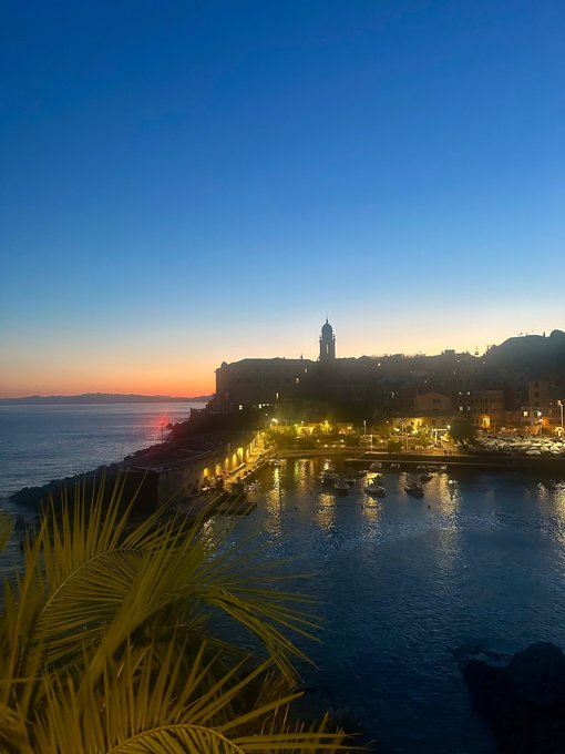 A scenic twilight view of a coastal town at dusk, showing a harbor with boats and illuminated buildings along the waterfront.