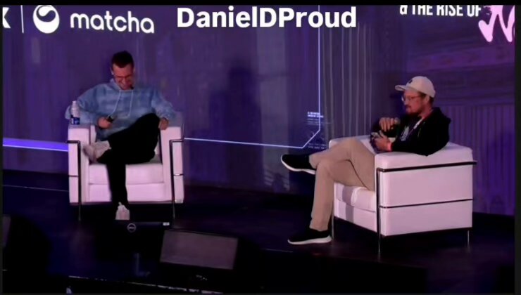 The image shows two men sitting in chairs at a stage or event, with one man wearing a blue shirt and the other wearing a white hat. The background features a purple wall with the words "DanielID Proud" written on it. There is also an image of a skateboard placed near the center of the scene. The focus of the image seems to be on the men sitting in chairs, possibly discussing or presenting something related to the event's theme or purpose.