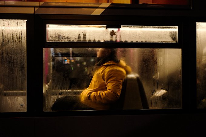 An atmospheric photograph of a person in a yellow jacket sitting inside what appears to be a train or tram, viewed through a foggy/rain-covered window with condensation.
