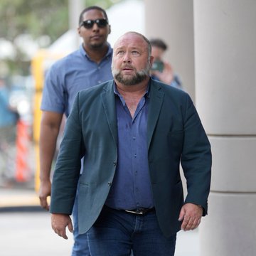 Outdoor photograph of a bearded man in a blue blazer walking forward, with a security guard wearing sunglasses visible behind him.