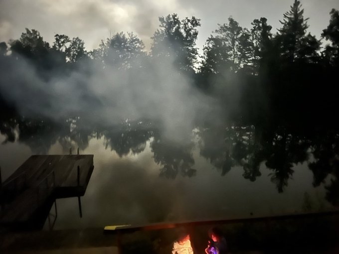 A moody, dark landscape shows a misty lake reflecting trees under a cloudy sky, with a wooden dock on the left and a small fire or light glowing in the foreground.
