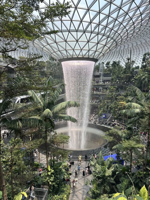 An interior view of a large indoor waterfall cascading from a glass dome ceiling, surrounded by lush tropical greenery and visitors below.