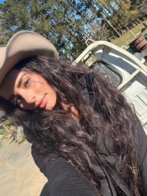 A woman with long, wavy dark hair and a tan beret takes a sunny outdoor selfie, with a white boat and tall trees visible in the background, radiating the cheerful, optimistic energy of a "good morning" greeting. Her relaxed, content expression mirrors the uplifting sentiment of wishing friends happiness and success to start the day.