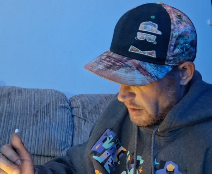 A man wearing a black graphic t-shirt and a black cap with a colorful brim featuring a cartoon design sits on a gray couch, looking down at something in his hands.
