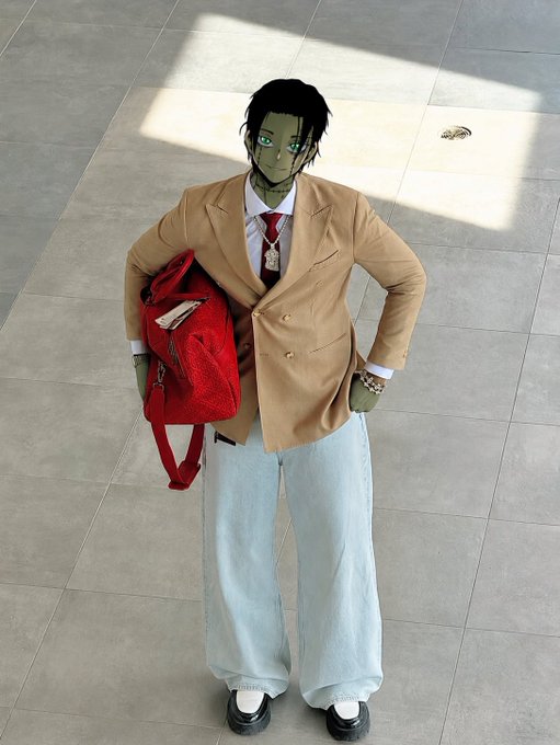 Person posing in a tan blazer, red tie, and light blue wide-leg pants, holding a red backpack, with a digitally altered cartoon face and green-tinted skin.