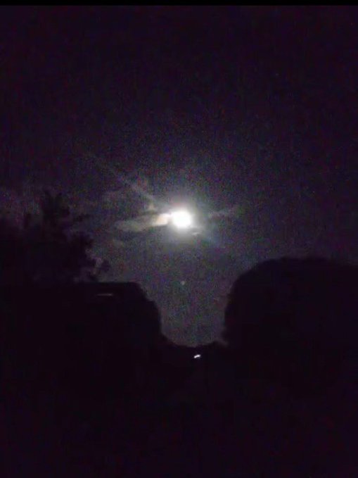 A nighttime photograph showing a bright full moon partially obscured by wispy clouds, with dark silhouettes of what appear to be a roof and tree branches in the foreground.